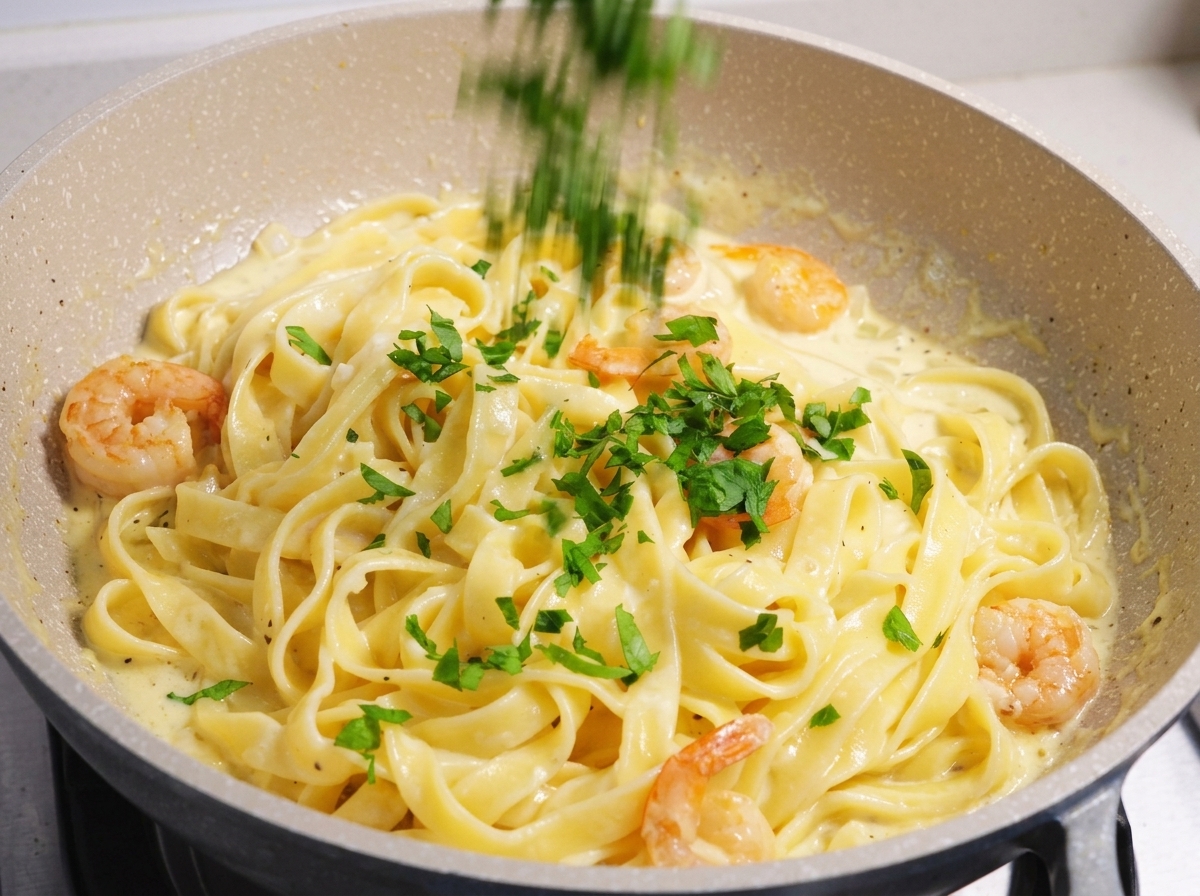 Frisch gehackte grüne Petersilie wird über ein cremiges Gericht mit Garnelen und Fettuccine-Pasta in einer hellen Pfanne gestreut.