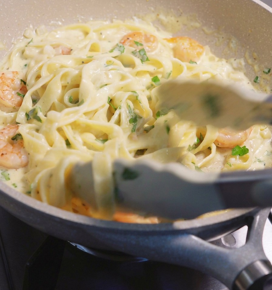 Mit einer Zange werden Fettuccine-Nudeln und rosa Garnelen in einer cremigen weißen Sauce in einer Pfanne angehoben und vermengt.