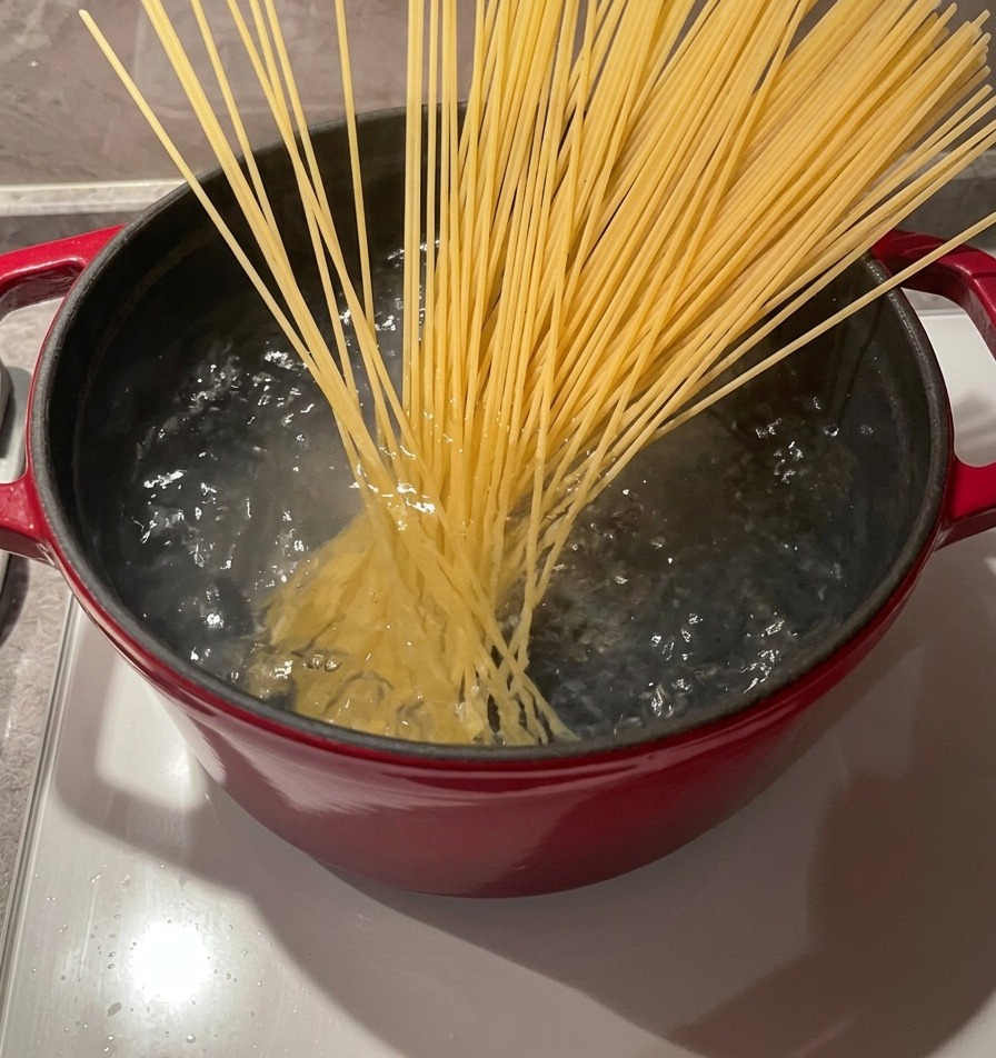 Ein Bündel trockener Spaghettinudeln wird mit Holzstäbchen in einen Topf mit sprudelnd kochendem Wasser gegeben.
