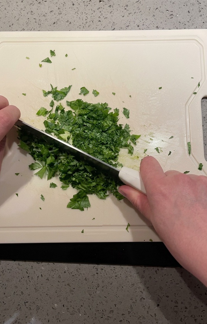 Vogelperspektive auf ein Messer, das einen Haufen frischer grüner Petersilie auf einem weißen Brett fein hackt.