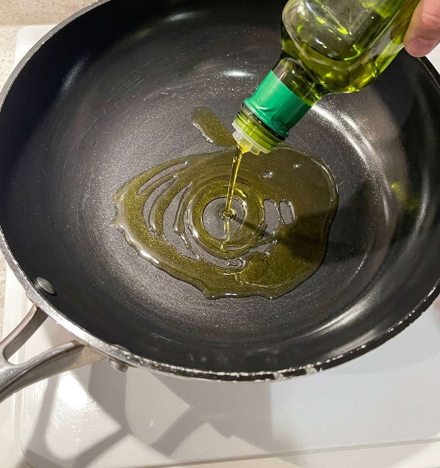 Goldenes Olivenöl wird aus einer Glasflasche in eine dunkle, beschichtete Pfanne gegossen.