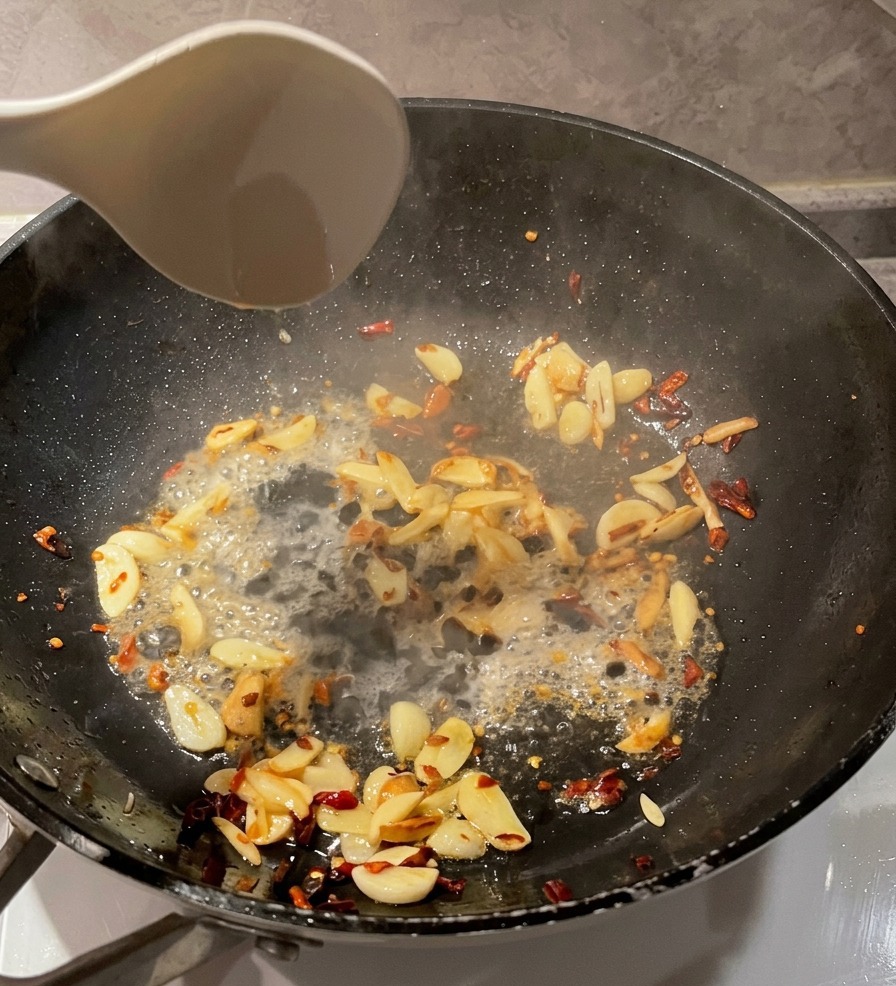 Eine weiße Kelle gießt klares Nudelwasser in eine schwarze Pfanne mit angebratenen Knoblauchscheiben und Chiliflocken.