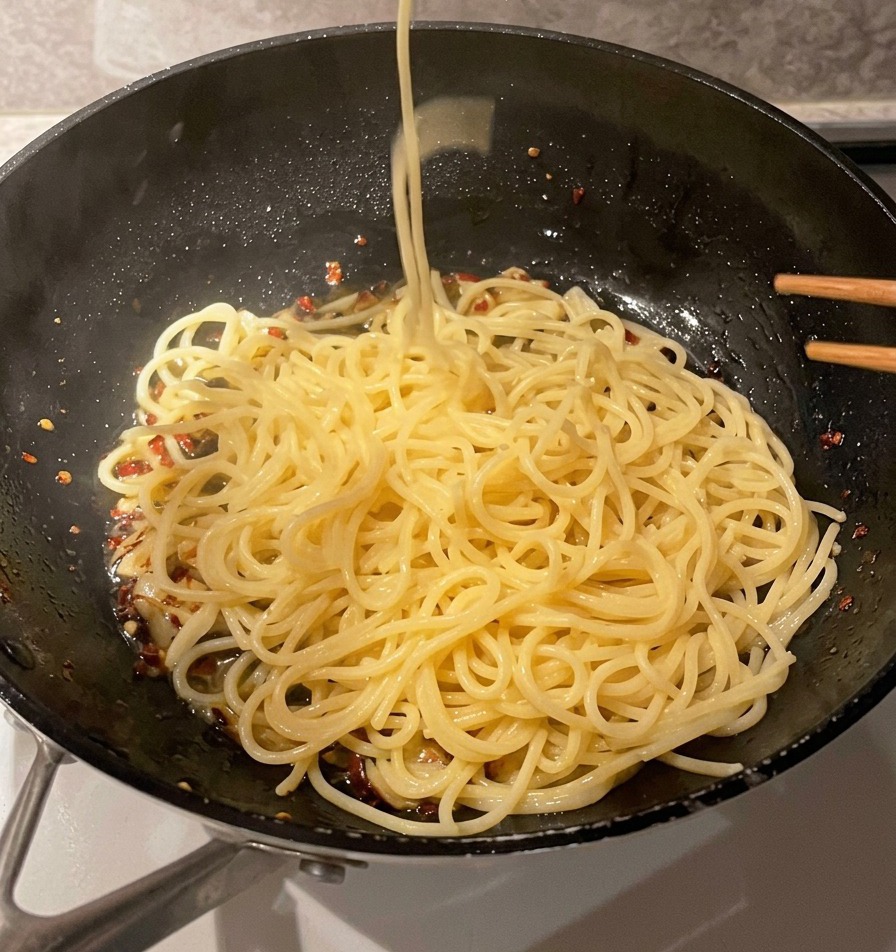 Ein Metallsieb gibt lange Stränge gekochter Spaghettinudeln in eine Pfanne mit Knoblauch und Öl.