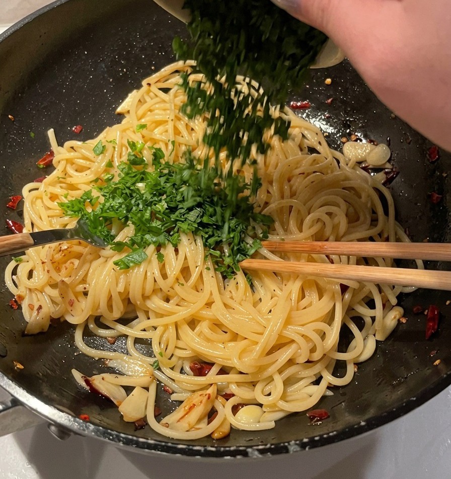 Eine Schüssel mit fein gehackter frischer Petersilie wird über einer Pfanne mit Knoblauch-Öl-Spaghetti geleert.