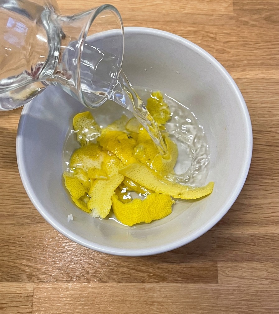 Klares Wasser wird aus einer Glaskaraffe in eine weiße Keramikschüssel gegossen, in der sich mehrere große Streifen frischer Zitronenschale befinden.