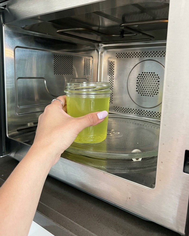 Eine Hand stellt ein Glas mit gelblich schimmerndem Zitronenwasser in eine moderne Mikrowelle aus Edelstahl.