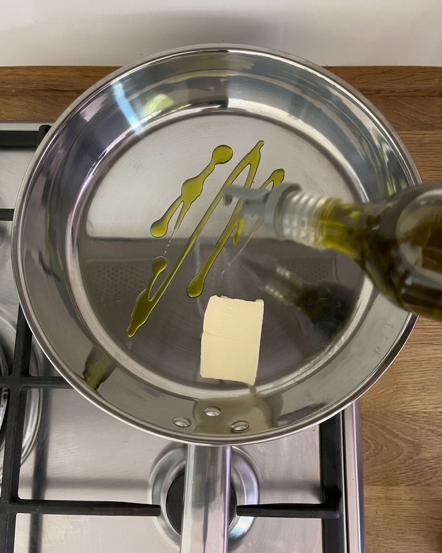 Olivenöl wird aus einer Flasche in eine glänzende Edelstahlpfanne gegossen, in der sich ein Stück Butter auf dem Herd befindet.