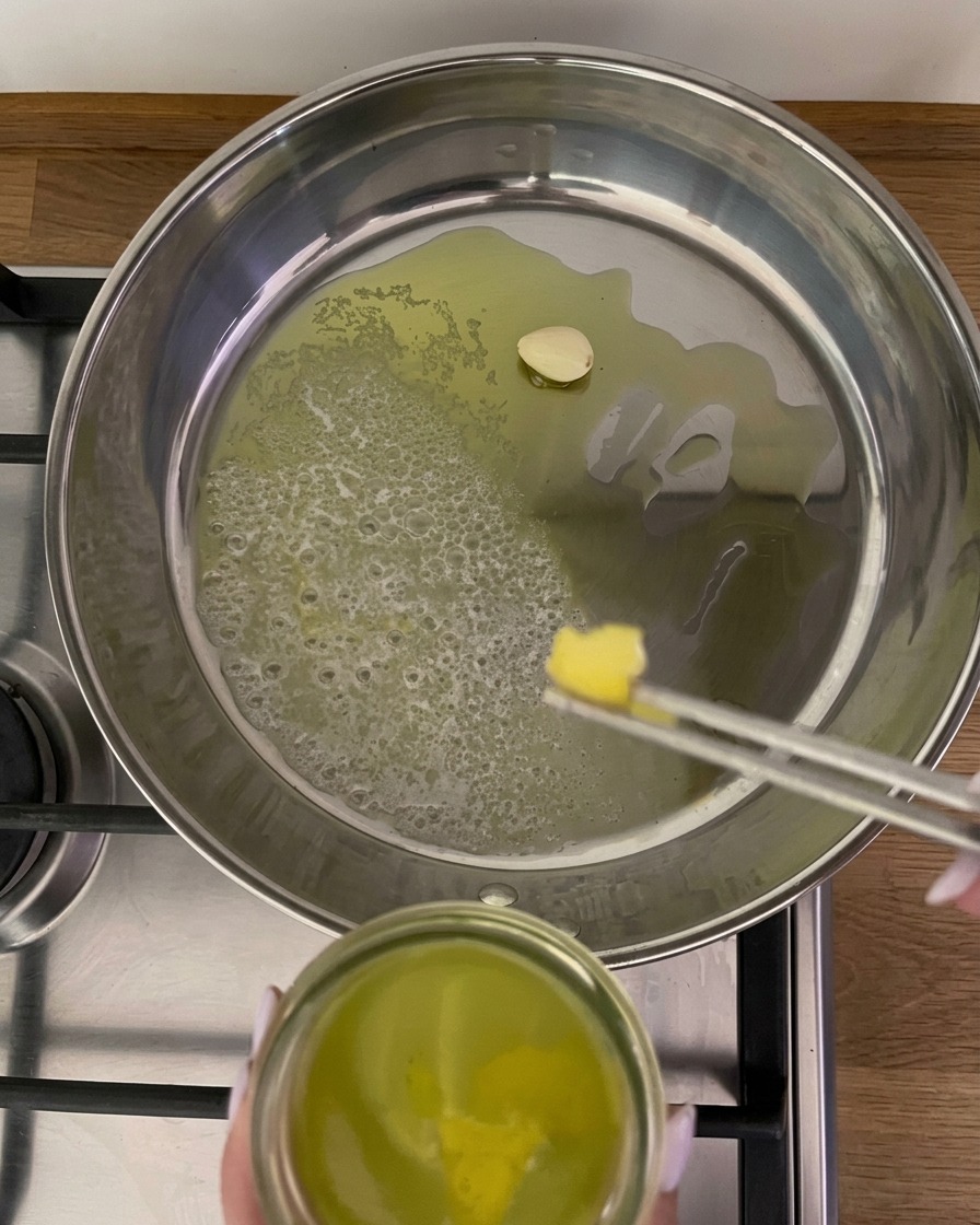 Eine Metallzange legt einen Streifen gelbe Zitronenschale in eine Pfanne mit schäumender Butter, Öl und einer einzelnen Knoblauchzehe.
