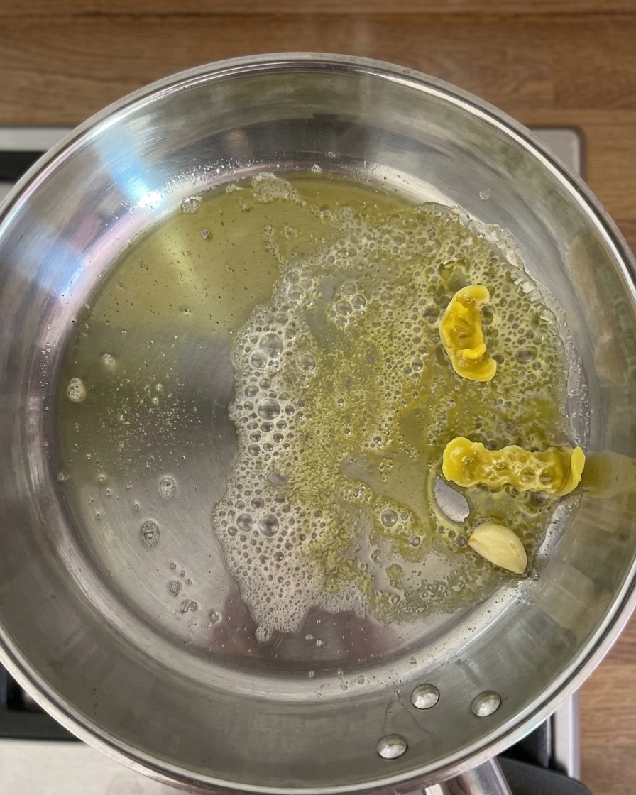 Warmes Zitronenwasser wird in eine Edelstahlpfanne mit angedünstetem Knoblauch und Zitronenschalen gegossen, wodurch Blasen in der Öl-Butter-Mischung entstehen.