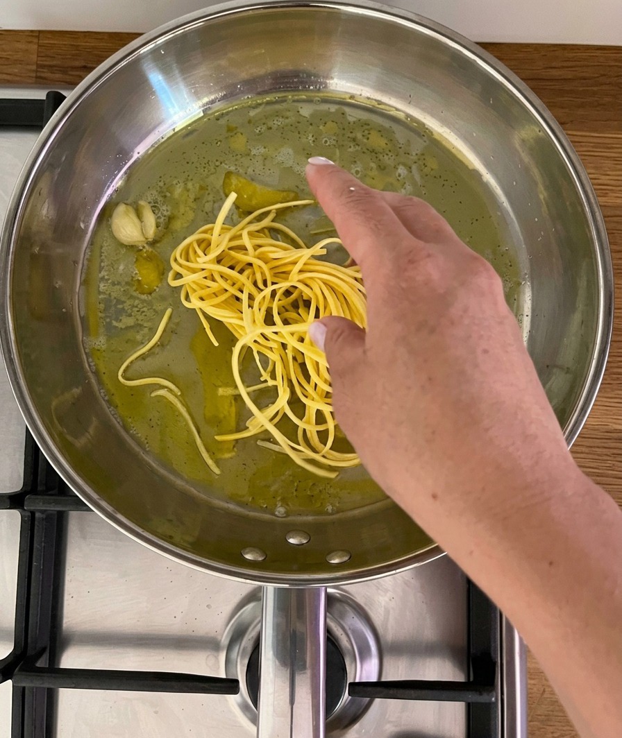 Eine Hand legt ein Nest aus frischen, gelben, spaghettiähnlichen Nudeln in eine Edelstahlpfanne mit Öl, einer Knoblauchzehe und Zitronenschale.