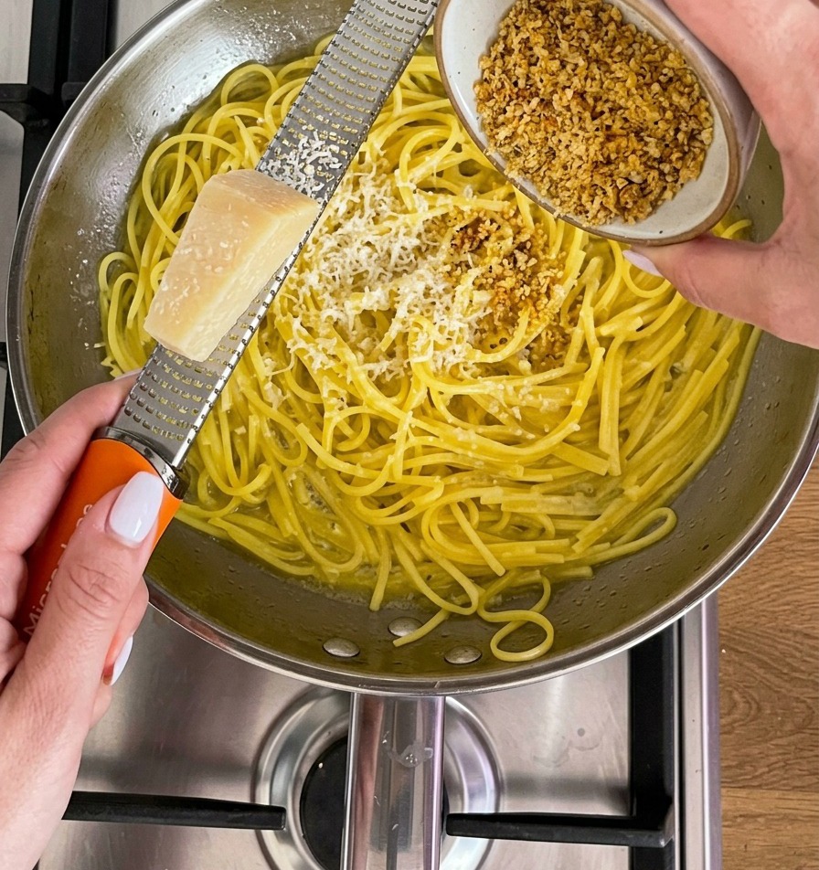 Ein Stück Parmesankäse wird mit einer Reibe direkt über einer Pfanne mit gelben Nudeln gerieben.