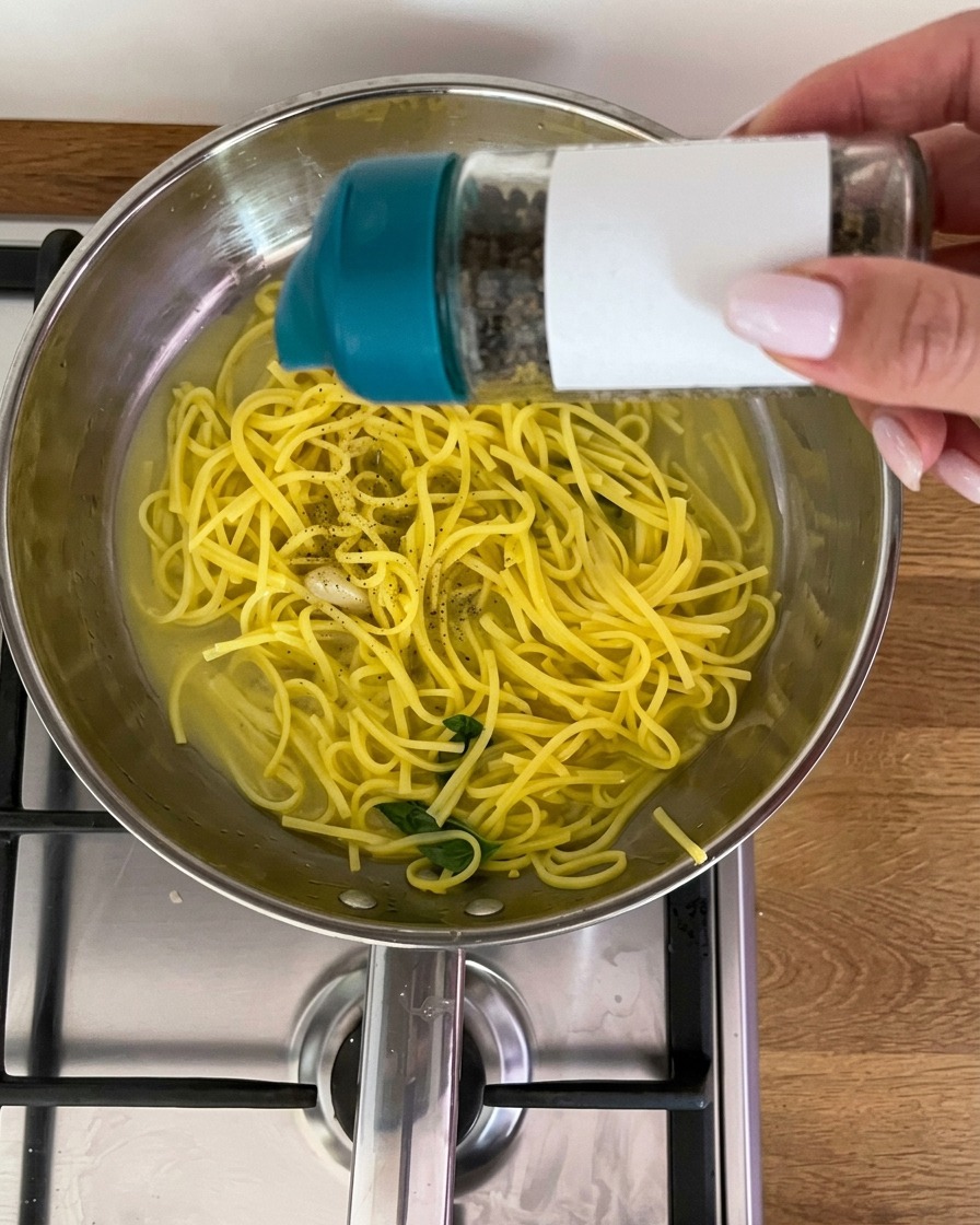 Frisch gemahlener schwarzer Pfeffer wird aus einer Mühle über eine Pfanne mit Zitronenpasta und frischen Basilikumblättern gestreut.
