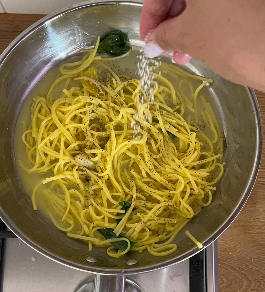 Nahaufnahme einer Hand, die eine Prise feines weißes Salz über eine Edelstahlpfanne mit gelben Spaghetti, frischem Basilikum und schwarzem Pfeffer streut.