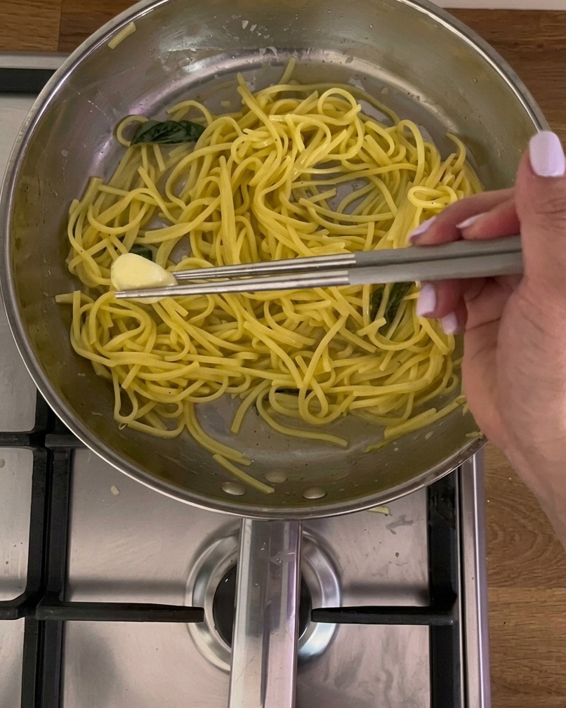 Metall-Essstäbchen nehmen eine weich gewordene Knoblauchzehe aus einer Edelstahlpfanne voller gelber Pasta und Basilikum.