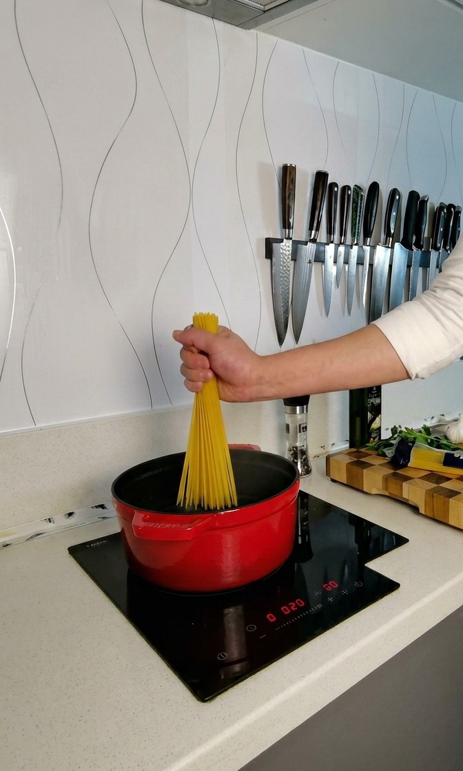 Eine Hand hält ein Bündel trockene Spaghetti über einen roten Topf mit kochendem Wasser und dreht die Nudeln auf, damit sie sich fächerförmig verteilen.