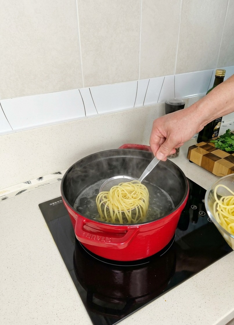 Ein Nest aus halbgekochten Spaghetti wird mit einem kleinen Schaumlöffel aus einem roten Topf mit dampfendem Wasser gehoben.