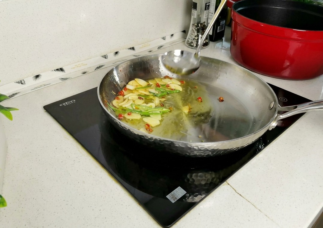 Eine Schöpfkelle gießt trübes Nudelwasser in eine Edelstahlpfanne mit angeschwitzten Knoblauchscheiben, Chilis und Petersilienstängeln.