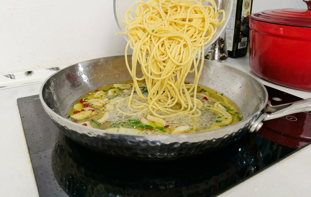 Gekochte Spaghetti werden aus einer Glasschüssel in eine Edelstahlpfanne mit einer köchelnden Knoblauch-Öl-Soßenbasis gegeben.