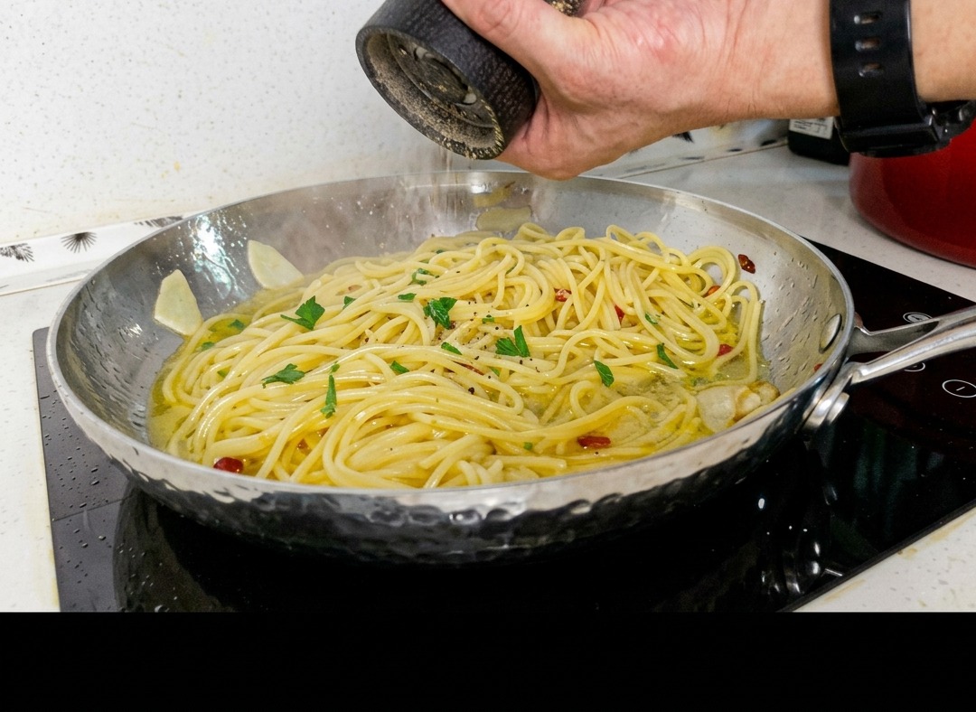 Ein Koch mahlt frischen schwarzen Pfeffer aus einer Mühle über eine Pfanne mit Spaghetti, die mit Knoblauch, Chili und Petersilie vermengt sind.