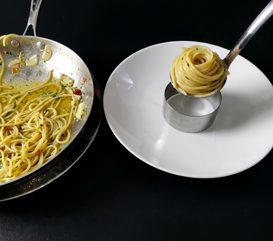 Nahaufnahme von Spaghetti, die mit einer Küchenpinzette und einer Schöpfkelle auf einem sauberen weißen Teller zu einem ordentlichen Zylinder in einem Metallring aufgedreht werden.