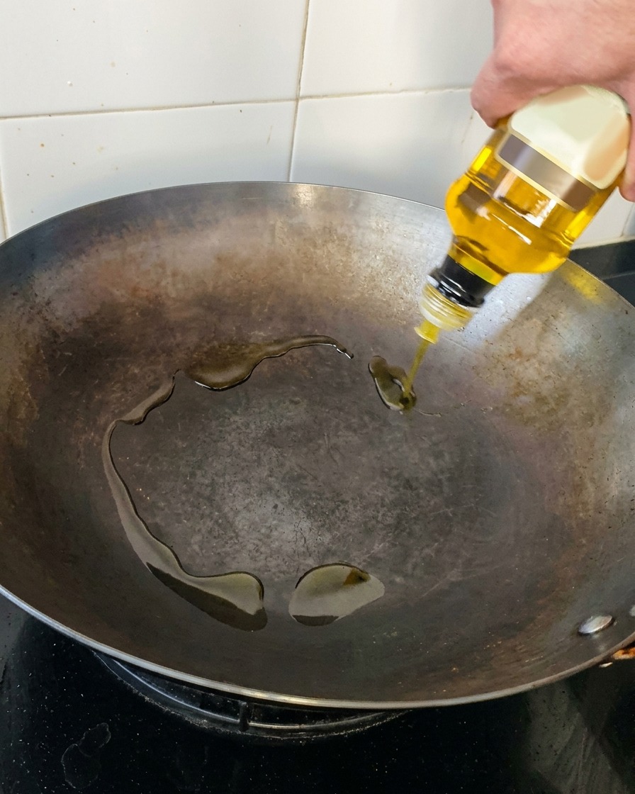 Eine Hand gießt goldenes Olivenöl aus einer klaren Glasflasche in einen dunklen, eingebrannten Wok aus Kohlenstoffstahl.