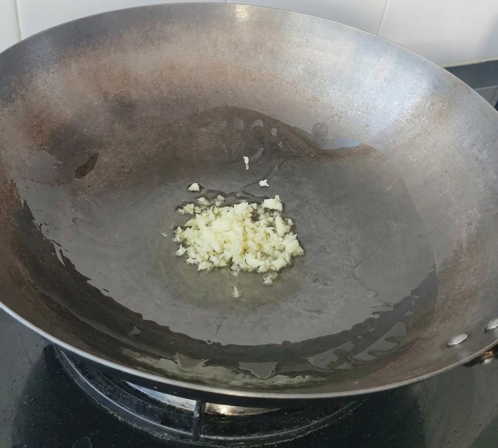 Gehackter Knoblauch brodelt in einer Pfütze Olivenöl auf dem Boden eines großen Woks aus Kohlenstoffstahl.