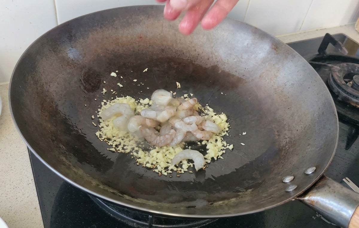 Eine Handvoll roher, glasiger Garnelen wird in einen heißen Wok mit angebratenem Knoblauch gegeben.
