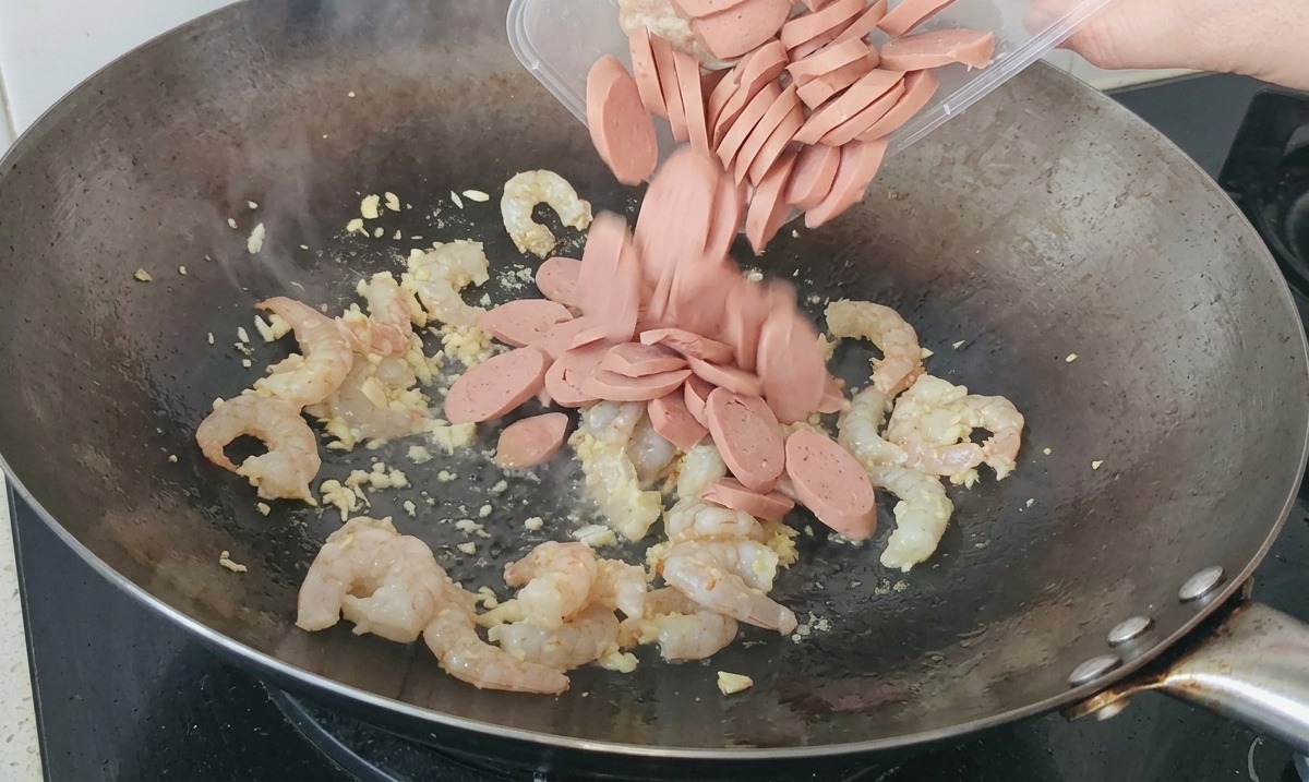 In scheiben geschnittene Würstchen werden aus einem Kunststoffbehälter in einen Wok mit bratenden Garnelen und Knoblauch geschüttet.