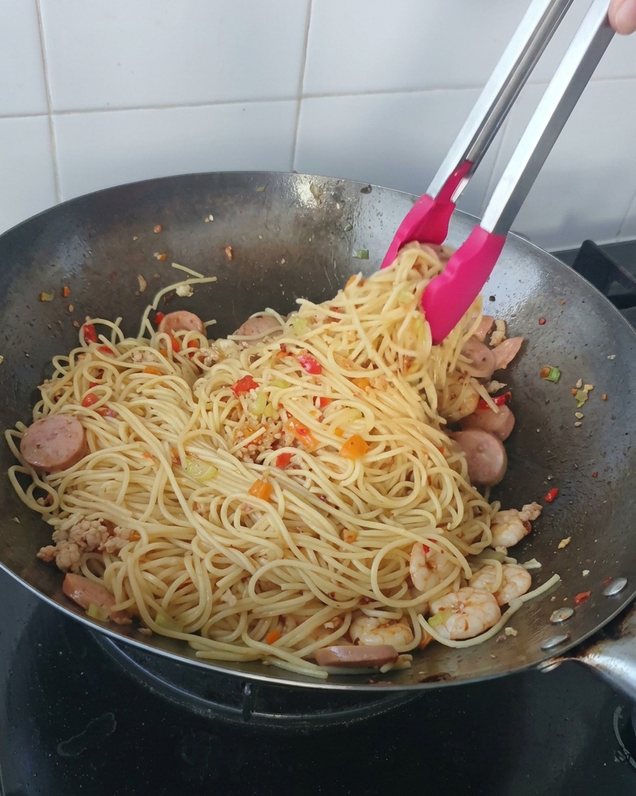 Eine Zange mit rosa Spitzen hebt eine große Portion Spaghetti, gemischt mit Garnelen und Wurstscheiben, aus einem Wok.