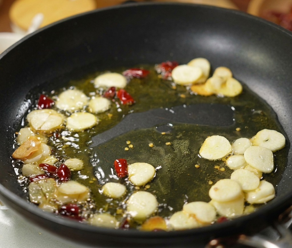 Dünne Knoblauchscheiben und getrocknete rote Chilistücke brodeln in etwas Olivenöl in einer schwarzen Bratpfanne.