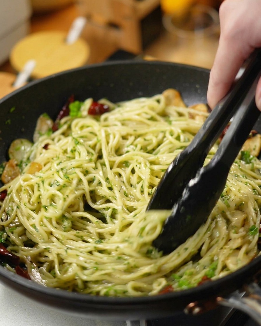 Schwarze Grillzange schwenkt Spaghetti, gehackte Petersilie, Knoblauch und Chilis in einer dunklen Pfanne.