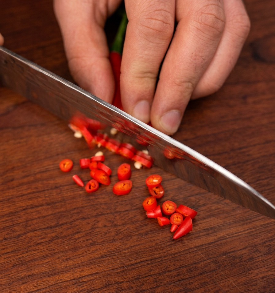 Ein scharfes Kochmesser schneidet kleine rote Birds-Eye-Chilis in dünne Ringe auf einem dunklen Holzschneidebrett.