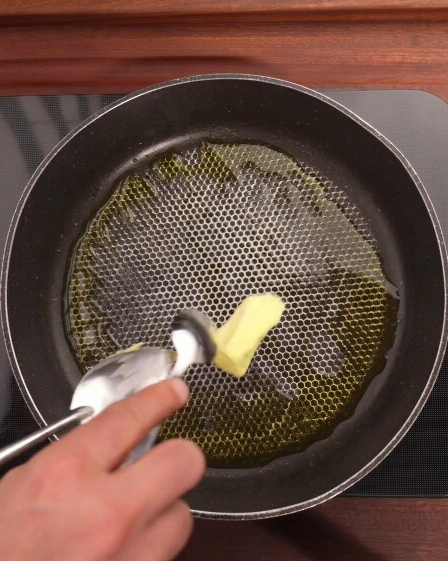 Eine Hand gibt ein Stück gelbe Butter mit einem Löffel in eine Pfanne mit Olivenöl, Knoblauchscheiben und Chili-Ringen.
