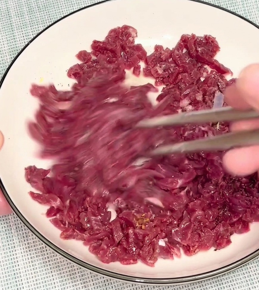 Eine Person vermengt rohes Rindfleisch in einer weißen Schüssel mit silbernen Essstäbchen, um die Marinade zu verteilen.