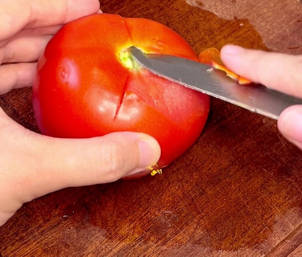 Hände ziehen vorsichtig mit einem Schälmesser die Haut von einer blanchierten Tomate ab.