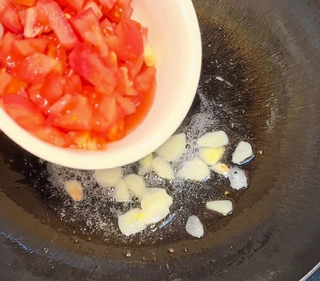 Gewürfelte Tomaten werden aus einer weißen Schüssel in eine Pfanne mit heißem Öl und goldbraunem Knoblauch gegeben.