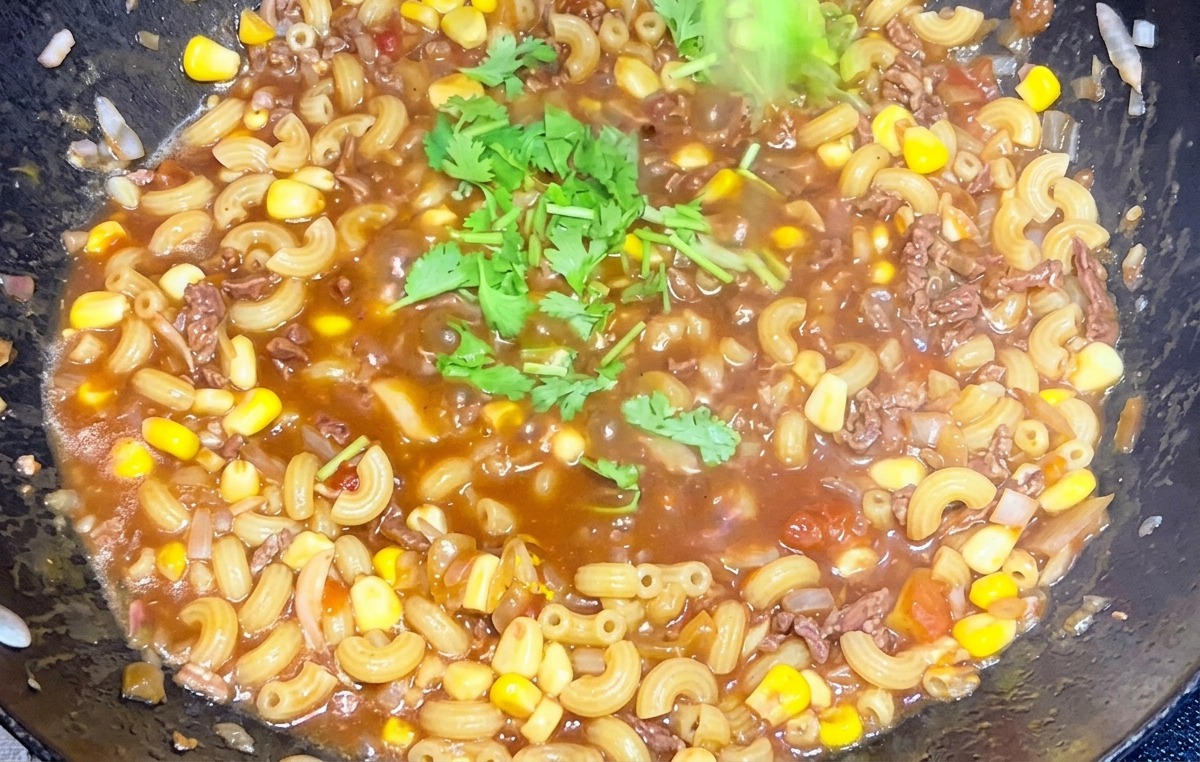 Eine Hand streut gehackten, grünen Koriander über eine dampfende Pfanne mit Rindfleisch-Tomaten-Makkaroni.
