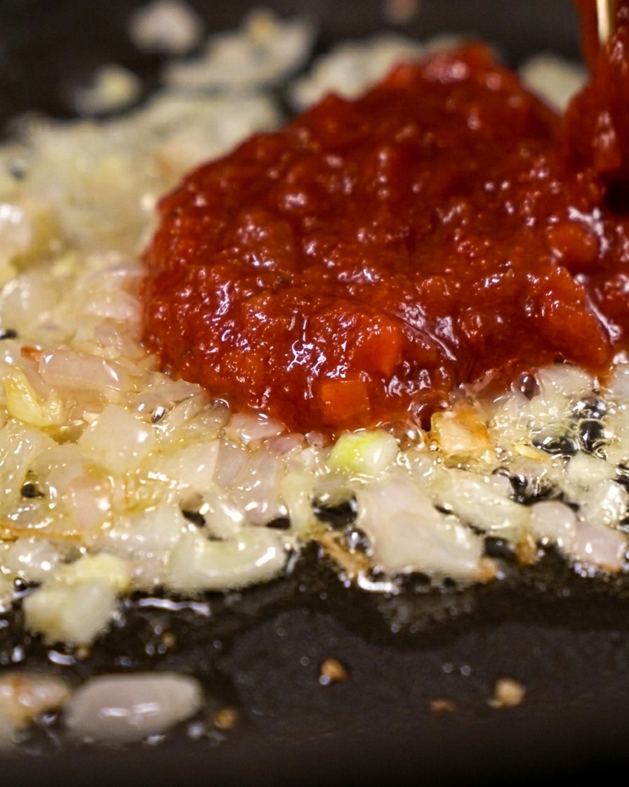 Ein großer Löffel mit dickflüssigem, tiefrotem Tomatenmark wird auf die brutzelnden, angeschwitzten Schalotten gegeben.