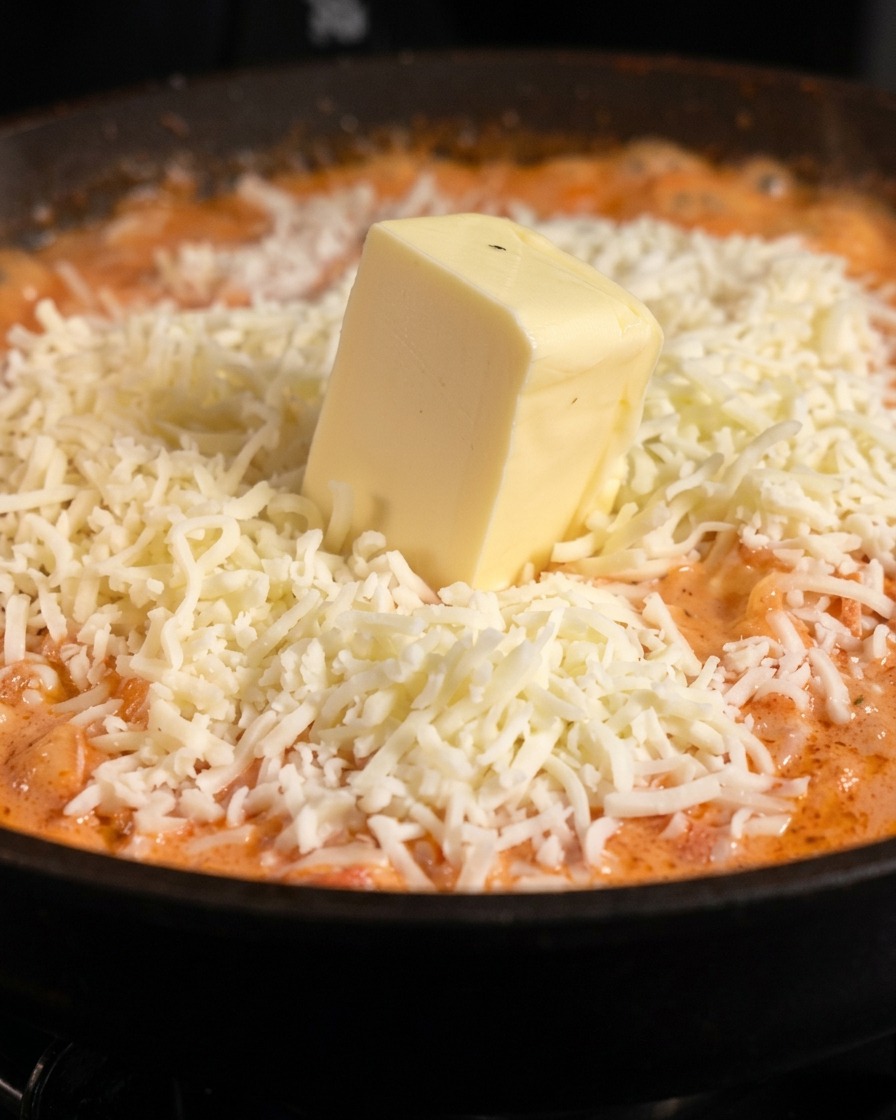 Ein dicker, quadratischer Block blasser Butter liegt auf einem Haufen geriebenem, weißem Mozzarella über orangefarbener Pastasauce.