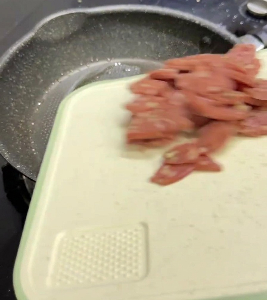Salami-Scheiben fallen von einem weißen Schneidebrett in eine dunkle, gesprenkelte Bratpfanne.