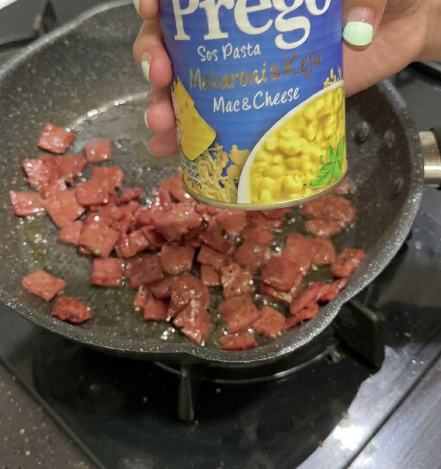 Eine Hand hält eine blau-gelbe Dose Prego Käsesauce über eine Pfanne mit angebratenen Salamiwürfeln.