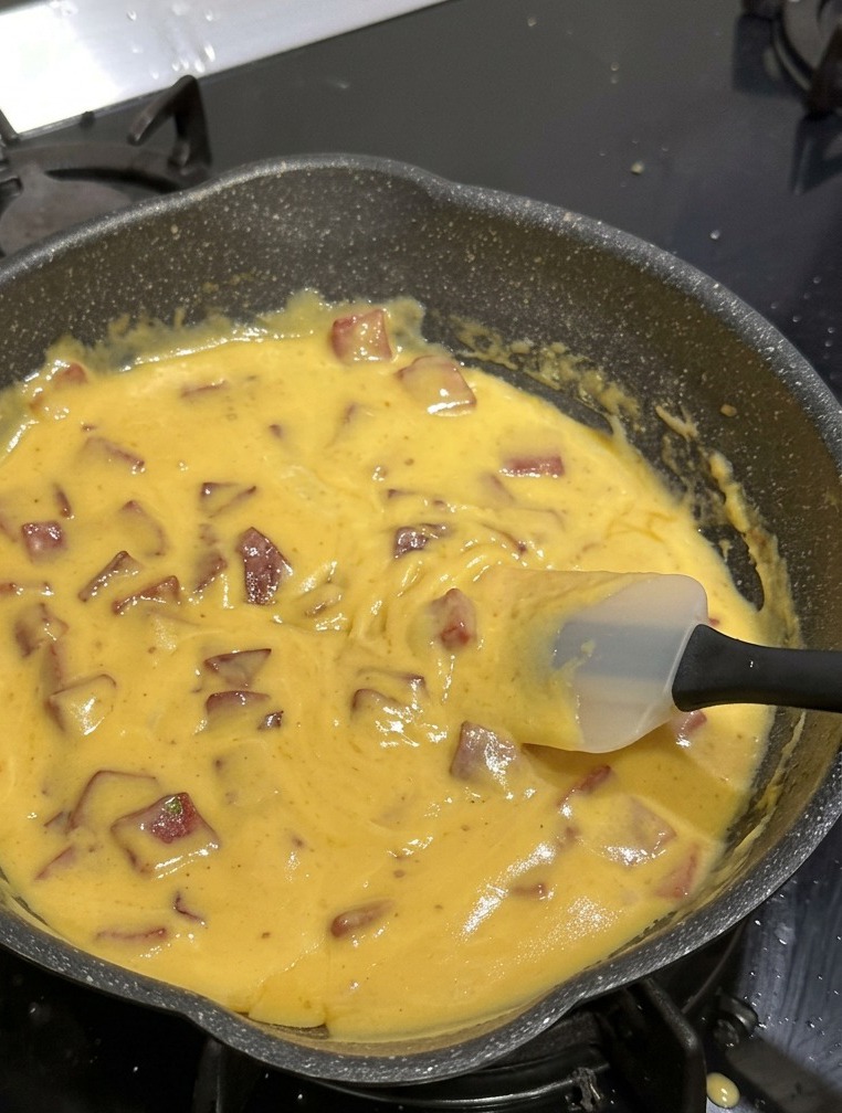 Eine schwarze, beschichtete Pfanne gefüllt mit Hörnchennudeln, Rindersalami und einer dicken, gelben Käsesauce wird mit einem weißen Silikonspatel umgerührt.