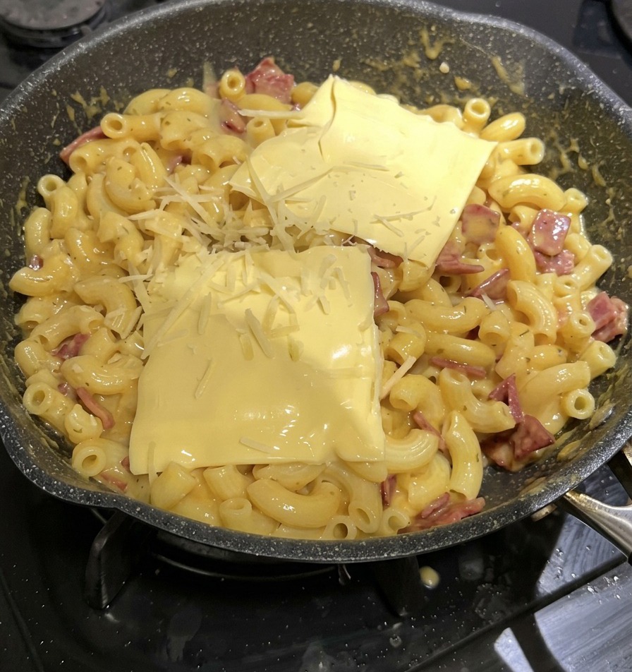 Zwei quadratische Schmelzkäsescheiben und geriebener Käse liegen auf den Mac and Cheese in der Pfanne.