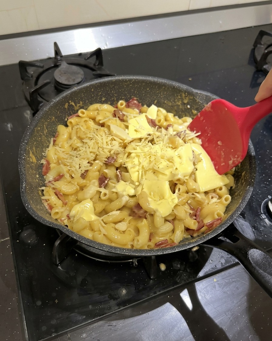 Hörnchennudeln mit Salami, Schmelzkäse und geriebenem Käse werden in einer schwarzen Pfanne mit einem roten Silikonspatel verrührt.