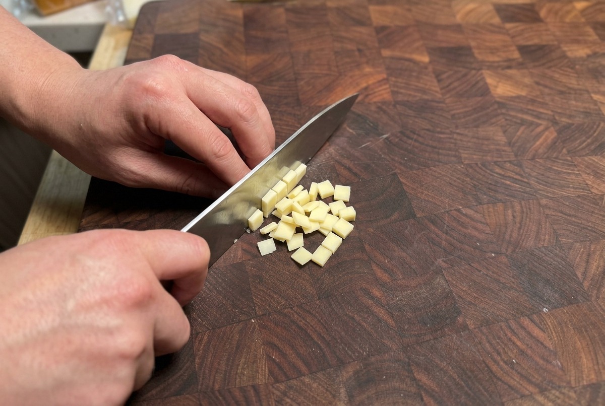 Hände schneiden mit einem Kochmesser ein Stück hellgelben Käse in kleine, gleichmäßige Würfel auf einem Holzschneidebrett.