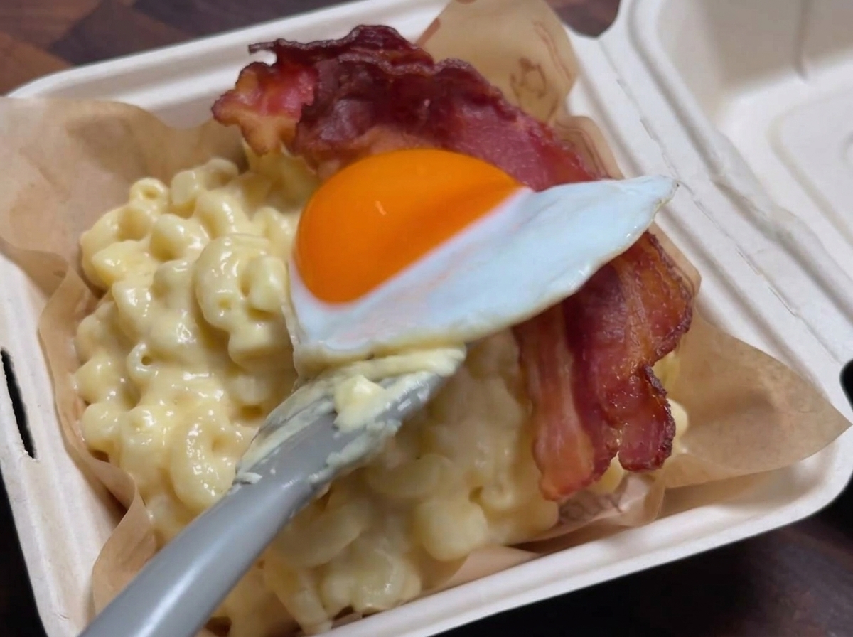 Ein Pfannenwender platziert vorsichtig ein Spiegelei auf einem Streifen knusprigem Speck über einer Portion cremiger Macaroni and Cheese.
