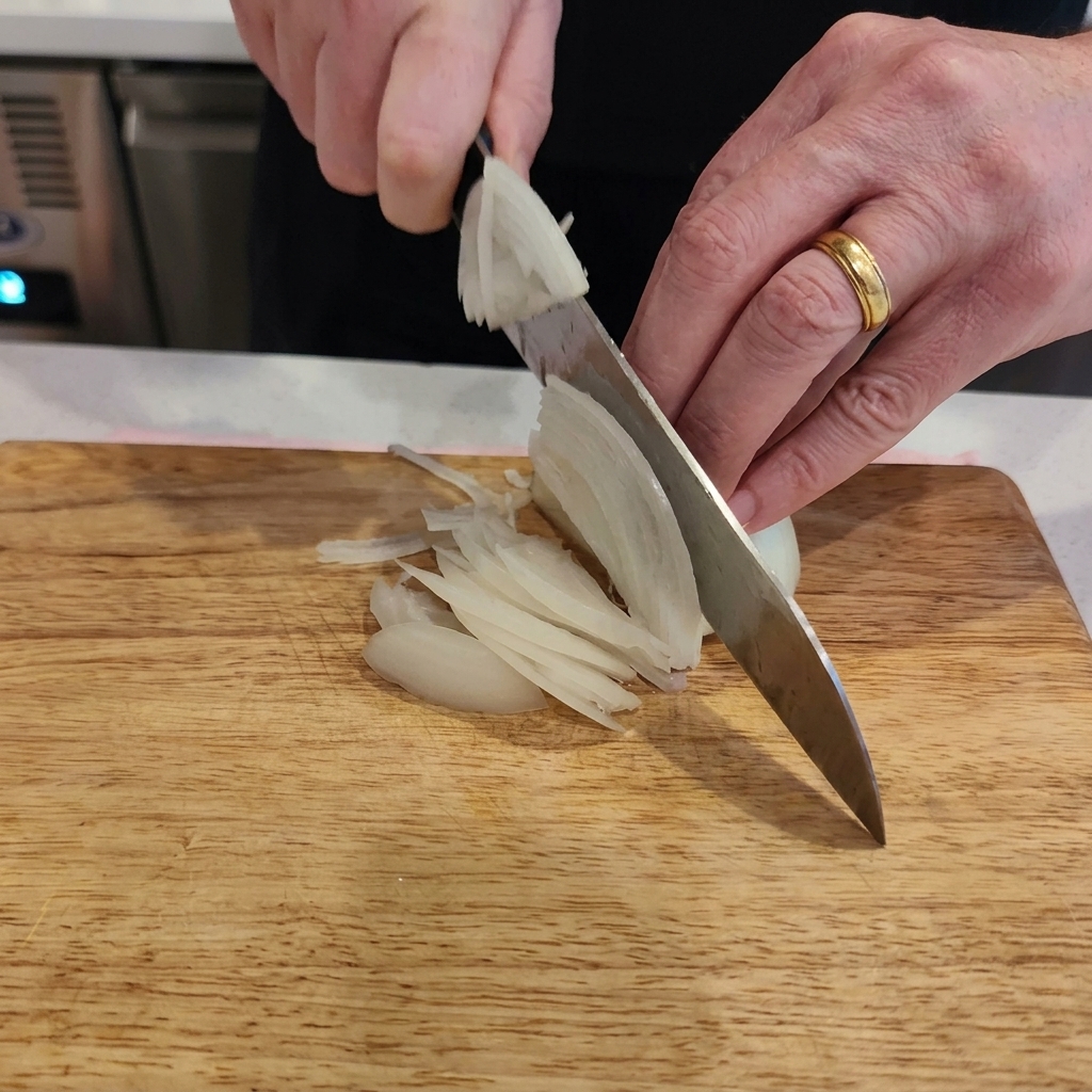 Eine Nahaufnahme von Händen, die eine weiße Zwiebel mit einem Kochmesser auf einem hellen Holzbrett in feine Streifen schneiden.