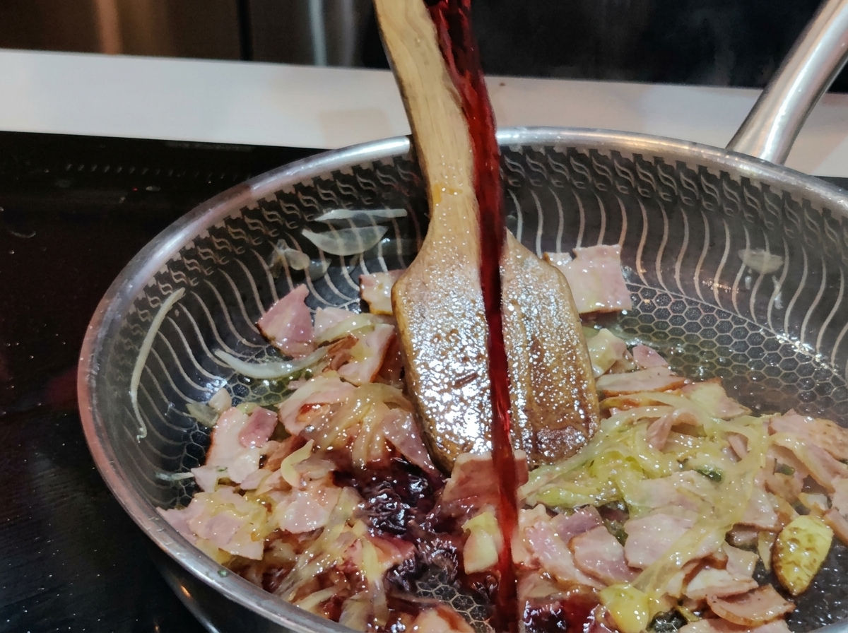 Ein kräftiger Schluck Rotwein wird aus einem Glas in eine Edelstahlpfanne mit brodelndem Speck und Zwiebeln gegossen.