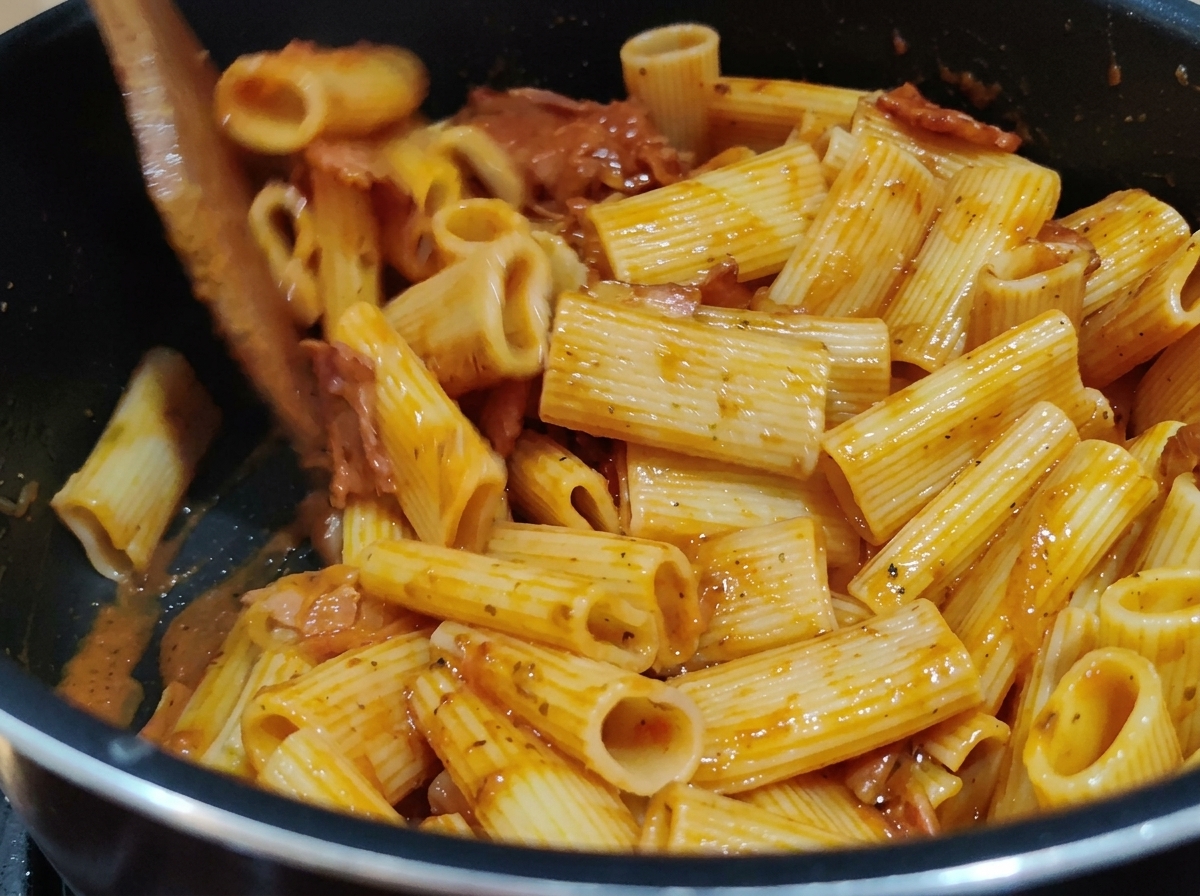 Rigatoni-Nudeln werden mit einem Holzspatel in einem Topf intensiv mit der reichhaltigen Tomatensauce vermengt.