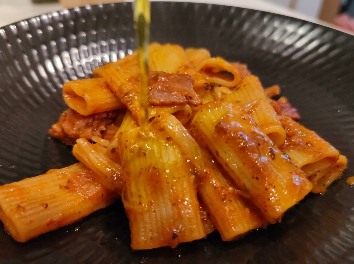 Ein feiner Strahl natives Olivenöl extra wird über eine angerichtete Portion Rigatoni Amatriciana gegossen.